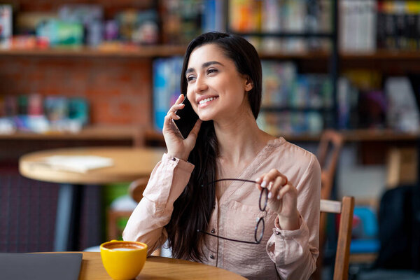 Attractive arab woman talking on smartphone while resting at cafe alone, looking aside and smiling, free space