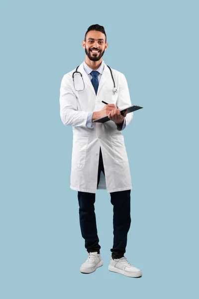 Счастливый врач-терапевт делает медицинские записи стоя над синим фоном — стоковое фото