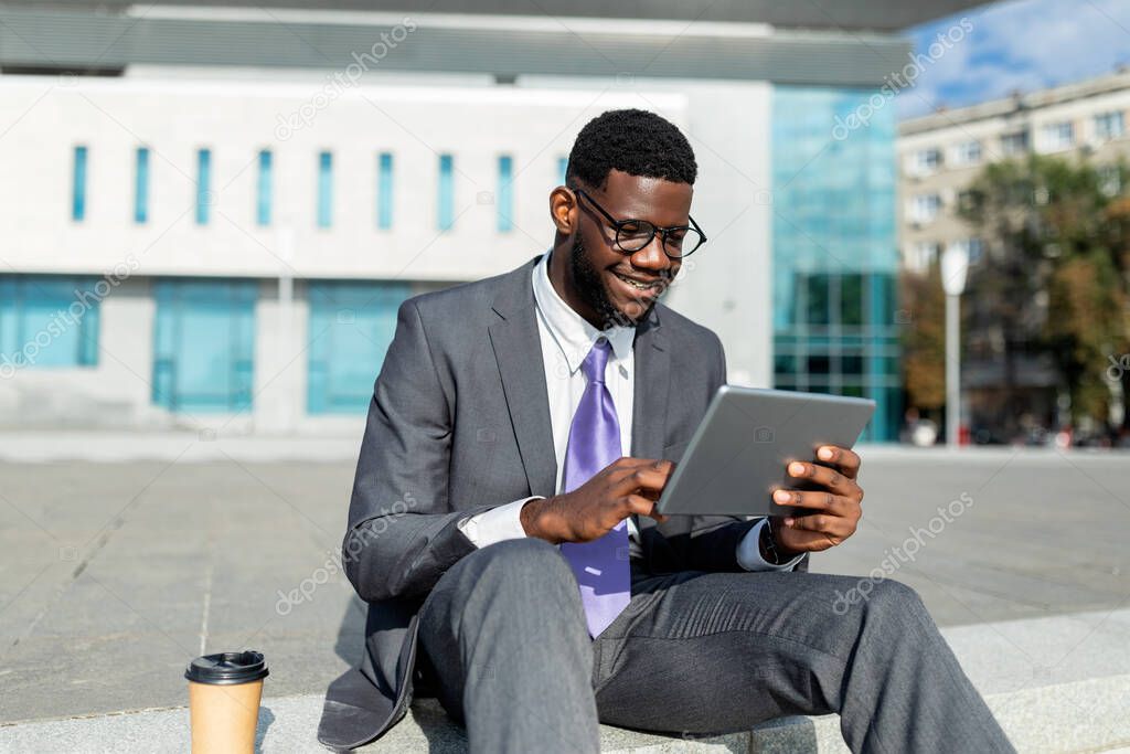 Happy african american insuranse agent working on tablet, browsing news on pad, sitting outdoors, free space