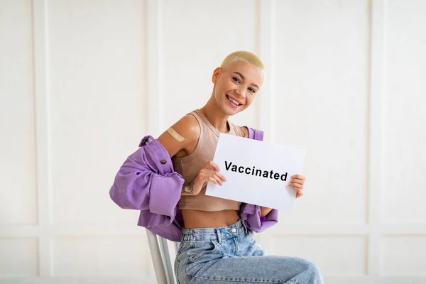 Vacunación con Covid-19. Señora feliz mostrando cartel con texto vacunado, sentado sobre la pared blanca y sonriendo a la cámara — Foto de Stock