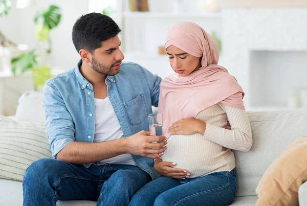 Péče a podpora během těhotenství. Muslim očekává, že žena se cítí špatně, muž dává jí sklenici vody, sedí na pohovce — Stock fotografie
