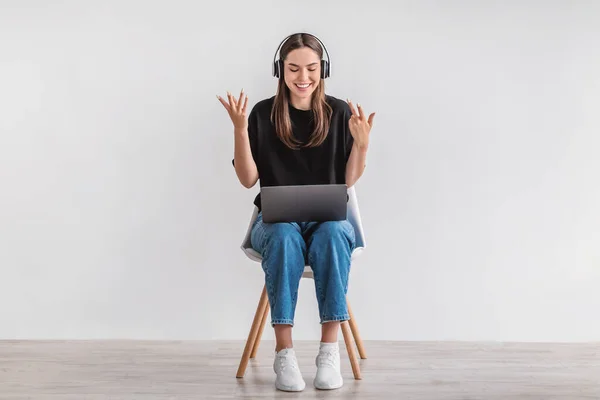 Девушка в наушниках делает видеозвонок на ноутбуке, сидит на стуле, участвует в удаленной конференции, чатится онлайн — стоковое фото