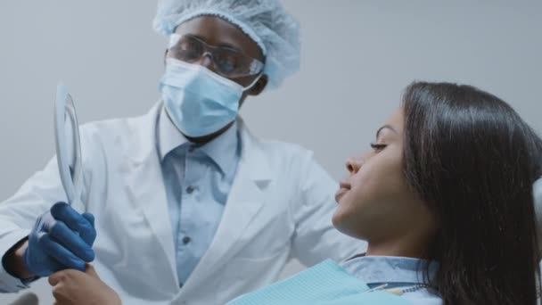 Profesionální muž zubař dávat zrcadlo černošky pacientky, vděčná žena těší výsledek léčby na klinice — Stock video