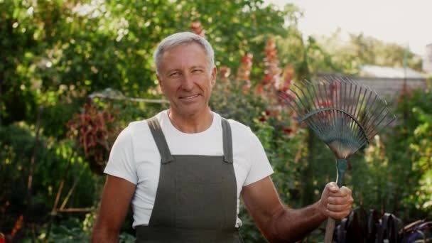 Snygg äldre man med lövkratta i handen Posing utomhus i trädgården — Stockvideo