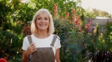 Bahçede elinde tırmıkla poz veren mutlu son sınıf güzeli kadın.