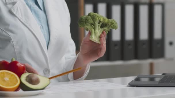 Genç profesyonel doktor beslenme uzmanı hastaya taze brokoli gösteriyor ve vitaminler hakkında konuşuyor, ofiste oturuyor. — Stok video