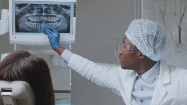 Чорний чоловік-стоматолог, який показує рентген щелепи на екрані пацієнту, консультує жінку про стан зубів і план лікування — стокове відео