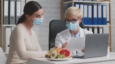 Genç bir kadın, profesyonel doktor diyetisyeniyle diyet planını tartışıyor. Ofiste koruyucu yüz maskeleri takıyor.