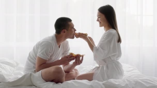Романтическая молодая пара, которая ест пиццу, расслабляясь в постели вместе — стоковое видео