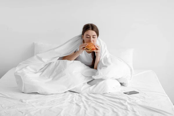 Smutna głodna młoda kobieta siedzi w domu na białym łóżku i je hamburgery, cierpiąc na depresję i problemy psychiczne — Zdjęcie stockowe
