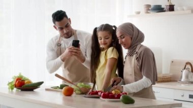 Orta Doğulu koca, evde eşi ve kızıyla yemek pişirirken fotoğraf çekiyor.