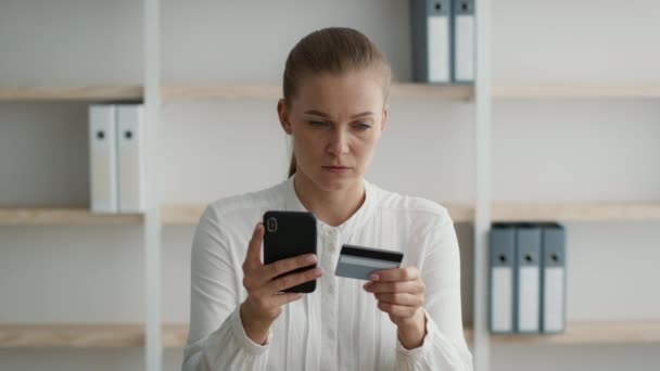 Empresaria dudosa con Smartphone y tarjeta de crédito haciendo compras en línea en el lugar de trabajo — Vídeos de Stock