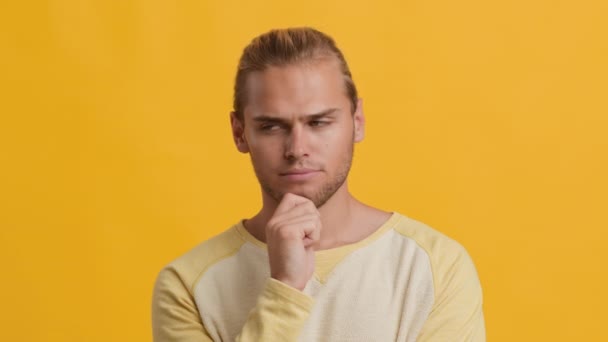 Töprengő fiatal szőke fickó megérinti az állát, miközben gondolkodik valamin, sárga háttér — Stock videók
