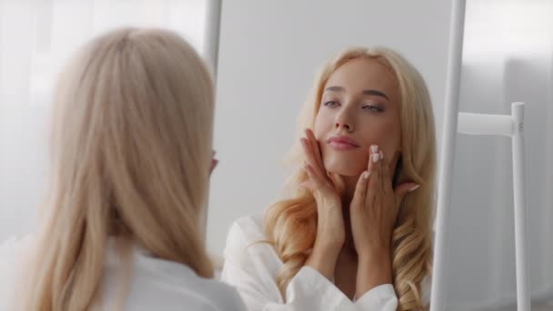 Массаж лица. Красивая блондинка смотрит на зеркало и трогает кожу — стоковое видео