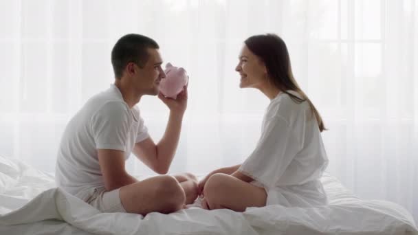 Jeune couple heureux assis au lit et secouant tirelire, vérification des économies — Video