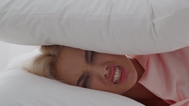 Rozdrażniona młoda blondynka zakrywająca głowę poduszką, rozdrażniona głośnym hałasem — Wideo stockowe