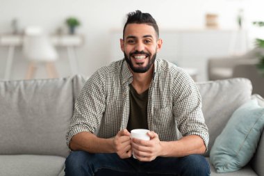 Genç gülümseyen Arap adam evdeki koltukta sıcak aromatik kahve içiyor, fotokopi çekiyor. Ilık içeceklerle yakışıklı Doğulu adam mola veriyor, oturma odasında tembellik ediyor.