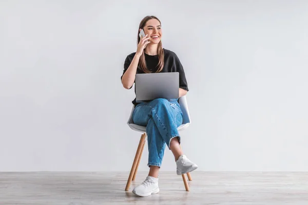 Ευτυχισμένη Καυκάσια κυρία με φορητό υπολογιστή που επικοινωνεί στο κινητό τηλέφωνο, κάθεται στην καρέκλα απέναντι σε λευκό τοίχο στούντιο — Φωτογραφία Αρχείου