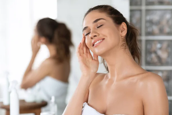 Vrouw met ogen gesloten aanbrengen crème op gezicht in de badkamer — Stockfoto