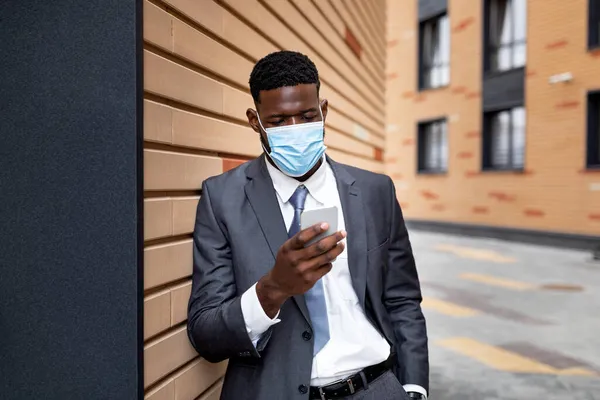 Empreendedor afro-americano em máscara protetora usando smartphone, de pé ao ar livre perto de prédio de escritórios — Fotografia de Stock