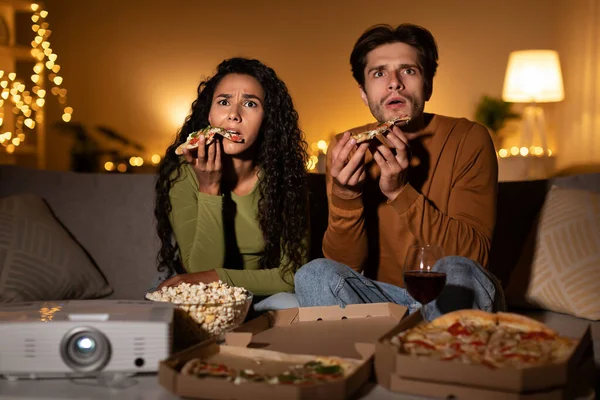 Shocked Couple Watching Horror Movie And Eating Pizza Using Home Cinema Projector Sitting On Sofa In Living Room Indoors At Night. Family Entertainment And Weekend Leisure Concept