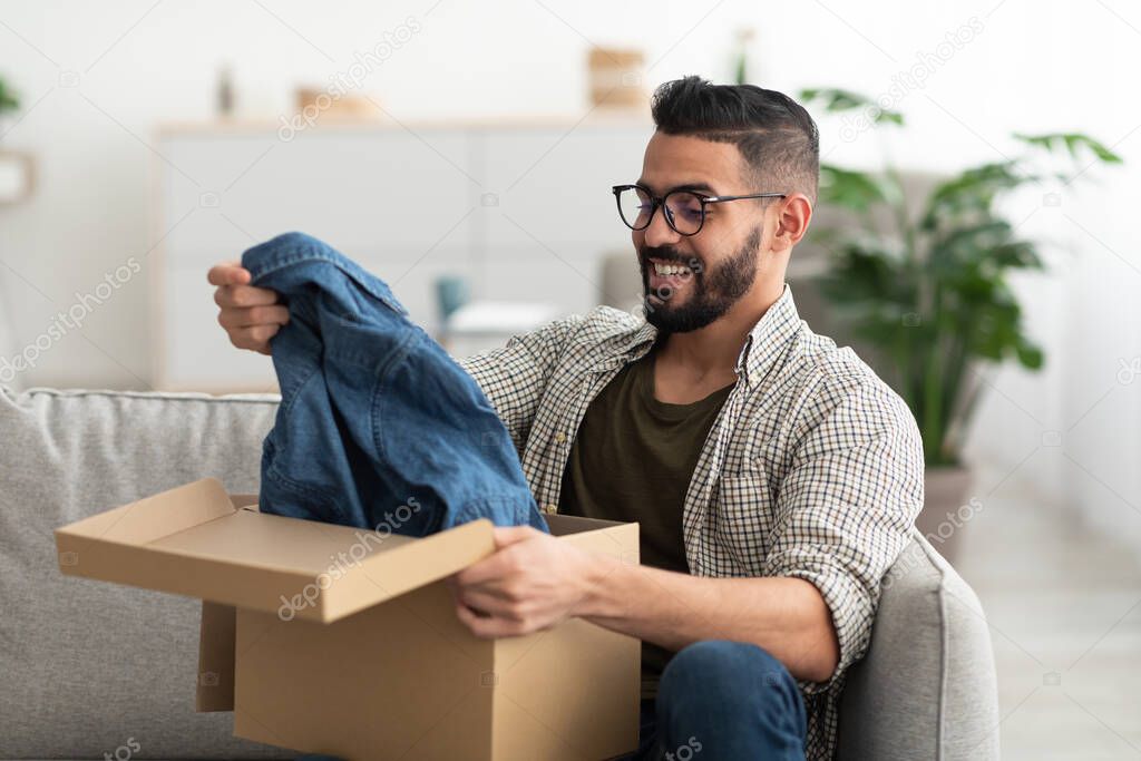 Satisfied Arab male client taking out new clothes from cardboard box, happy with delivery service at home. Millennial Eastern guy unpacking cardboard parcel, receiving online store order, indoors