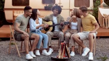 Neşeli çok ırklı arkadaşlar kamp ateşi etrafında oturur, gitar çalar ve şarkı söylerler.