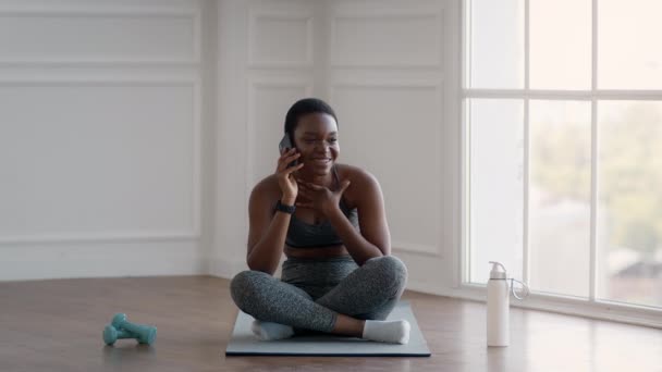 Спортивная молодая черная женщина разговаривает по мобильному телефону, расслабляясь после домашней тренировки — стоковое видео