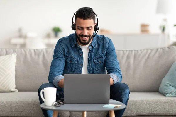 Práce na volné noze. Mladý usměvavý Arab pracuje na notebooku z domova, nosí sluchátka, používá PC počítač — Stock fotografie