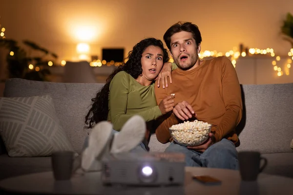 Pareja asustada viendo película de terror en shock usando proyector en interiores —  Fotos de Stock