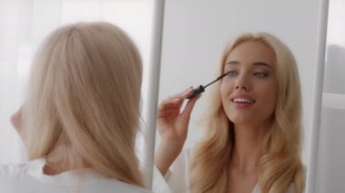 Günlük makyaj. Genç Güzel Sarışın Kadın Aynaya Bakıyor ve Maskarayı Uygulıyor