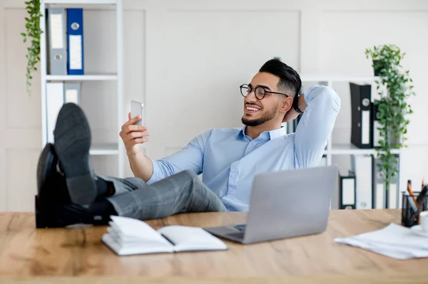 Happy young Arab man in office wear using smartphone, having break, putting feet on table at workplace with laptop