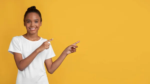 Улыбающаяся чернокожая девочка-подросток в белой футболке, указывающая пальцами в сторону свободного пространства — стоковое фото