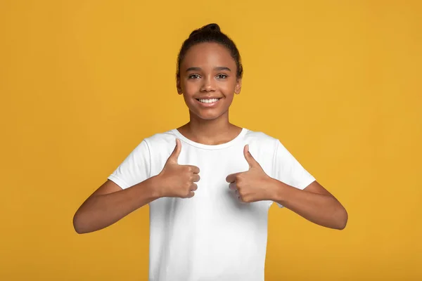 Smiling young afro american girl in white t-shirt rejoices to success and shows thumbs up — Stock Photo, Image