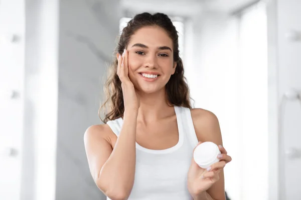 Headshot de alegre feminino hidratante rosto aplicando creme no banheiro — Fotografia de Stock