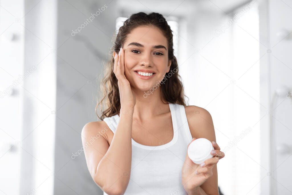 Headshot Of Joyful Female Moisturizing Face Applying Cream In Bathroom