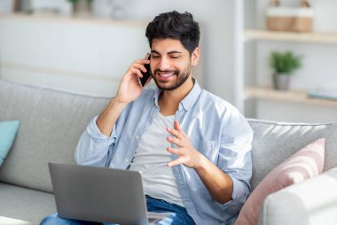 Heyecanlı Arap adam dizüstü bilgisayar kullanıyor ve akıllı telefonla konuşuyor oturma odasındaki koltukta otururken, boş yer