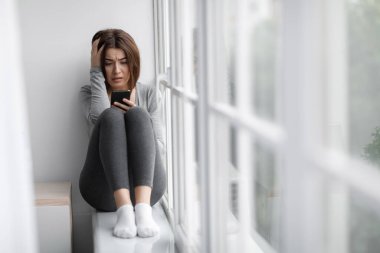 Kafası karışık üzgün Avrupalı bin yıllık bayan pencere eşiğinde oturur ve akıllı telefondan mesaj okur.