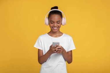 Beyaz tişörtlü, kulaklıklı, telefonda video izleyen gülümseyen siyah kız.