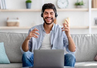 Uzaktan iletişim konsepti. Kulaklıklı, dizüstü bilgisayarlı kameraya el kol hareketi yapan mutlu bin yıllık Arap adam.