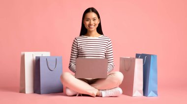 Çevrimiçi satış konsepti. Dizüstü bilgisayarlı neşeli Asyalı kadın alışveriş torbalarının arasında oturuyor.