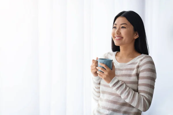 Ontspannen Koreaanse vrouw drinken koffie terwijl staande in de buurt van venster thuis — Stockfoto