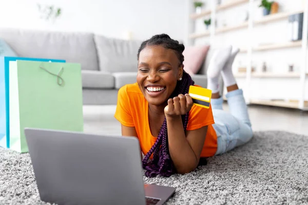 Ung afroamerikansk kvinna köper online med kreditkort och bärbar dator, ligger på golvet hemma — Stockfoto