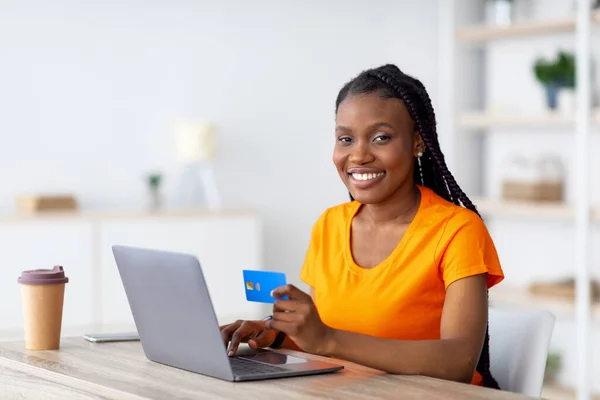 Cheerful black woman with laptop and credit card sitting at desk, shopping online, making remote payment at home