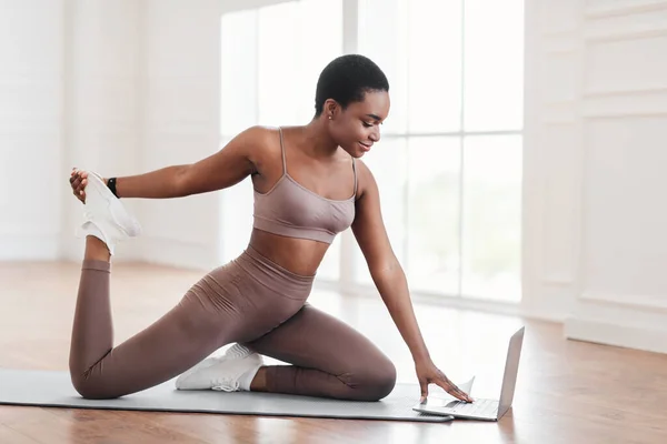 Весела чорна жінка розтягує м'язи ніг за допомогою ноутбука — стокове фото