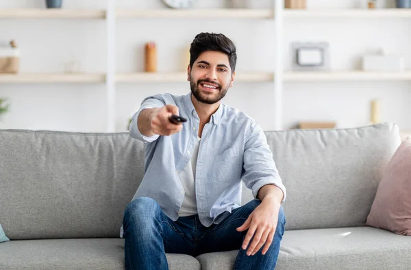 Smiling arab guy watching comedy on TV at home, sitting on sofa in living room, holding TV remote, copy space