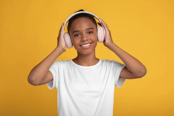 Alegre bonita adolescente afro-americana menina no branco t-shirt no fones de ouvido ouvir música, isolado no fundo amarelo — Fotografia de Stock