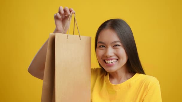 Glückliche koreanische Shopaholic zeigt Papier-Einkaufstasche, gelber Hintergrund — Stockvideo