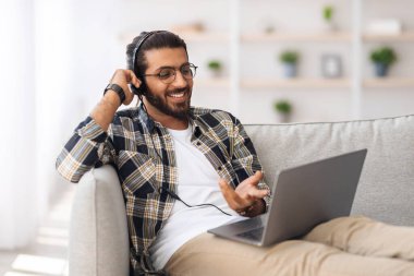 Kanepede oturan neşeli Hintli adam, dizüstü bilgisayar ve kulaklık kullanıyor.