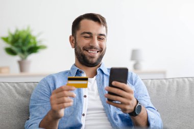 Sakallı, mutlu, yetişkin bir beyaz adam kredi kartı tutuyor ve internet üzerinden akıllı telefona para ödüyor.